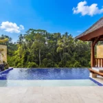 A private infinity pool at a boutique hotel bali with a stone relief carving and a traditional wooden gazebo, overlooking a bright green jungle under a blue sky.