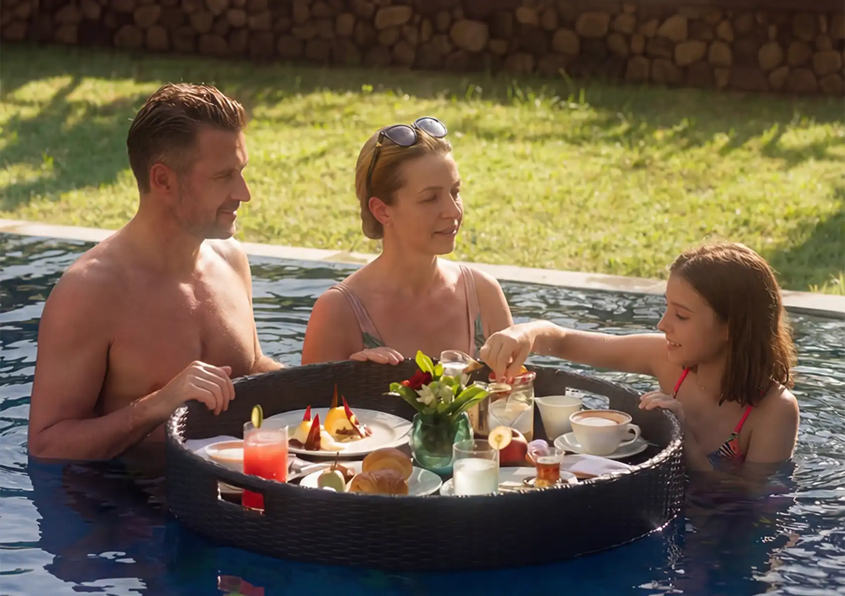 A family of three enjoying a floating breakfast in a swimming pool at a boutique hotel bali, with a woven tray filled with food and drinks