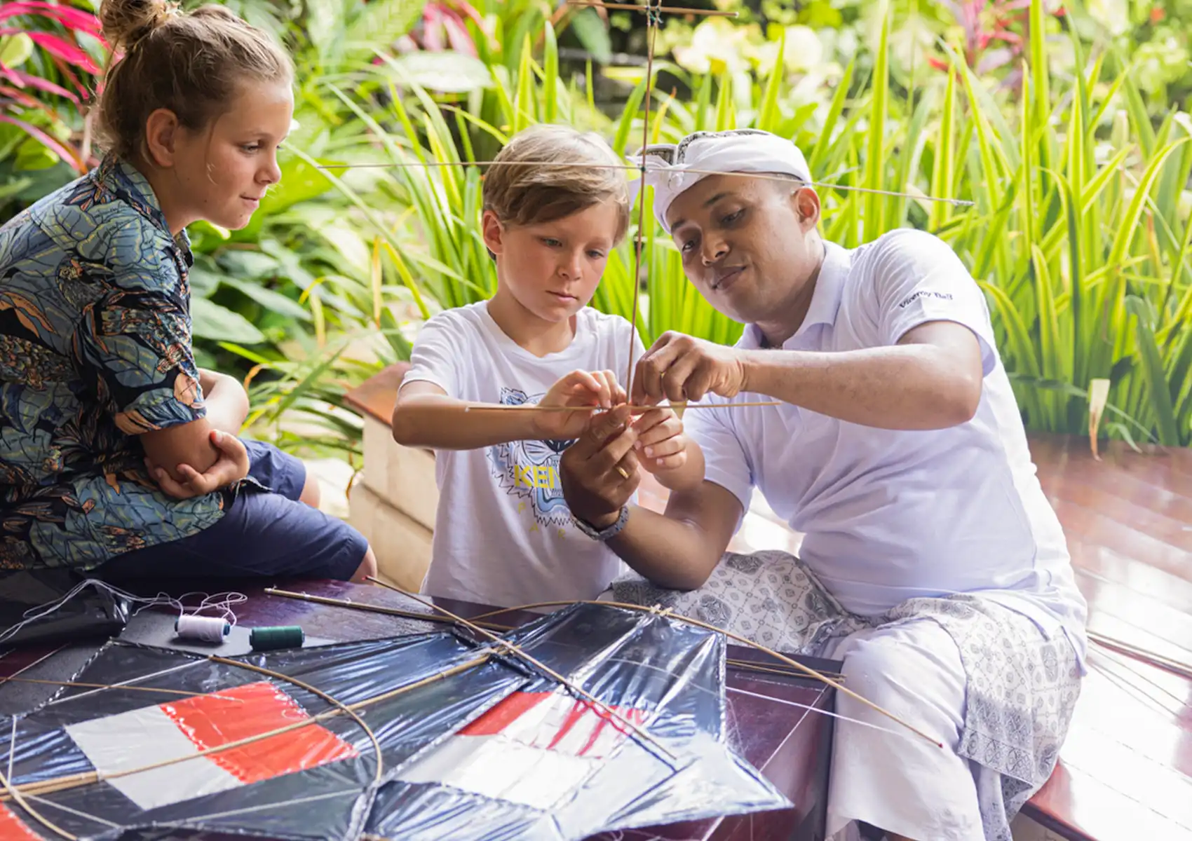 Two children and a local staff member building a traditional kite together on a wooden deck at a boutique hotel bali.
