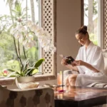 A smiling woman wearing a white bathrobe reading a brochure while sitting in a comfortable armchair next to a large potted white orchid and a window at a boutique hotel bali.