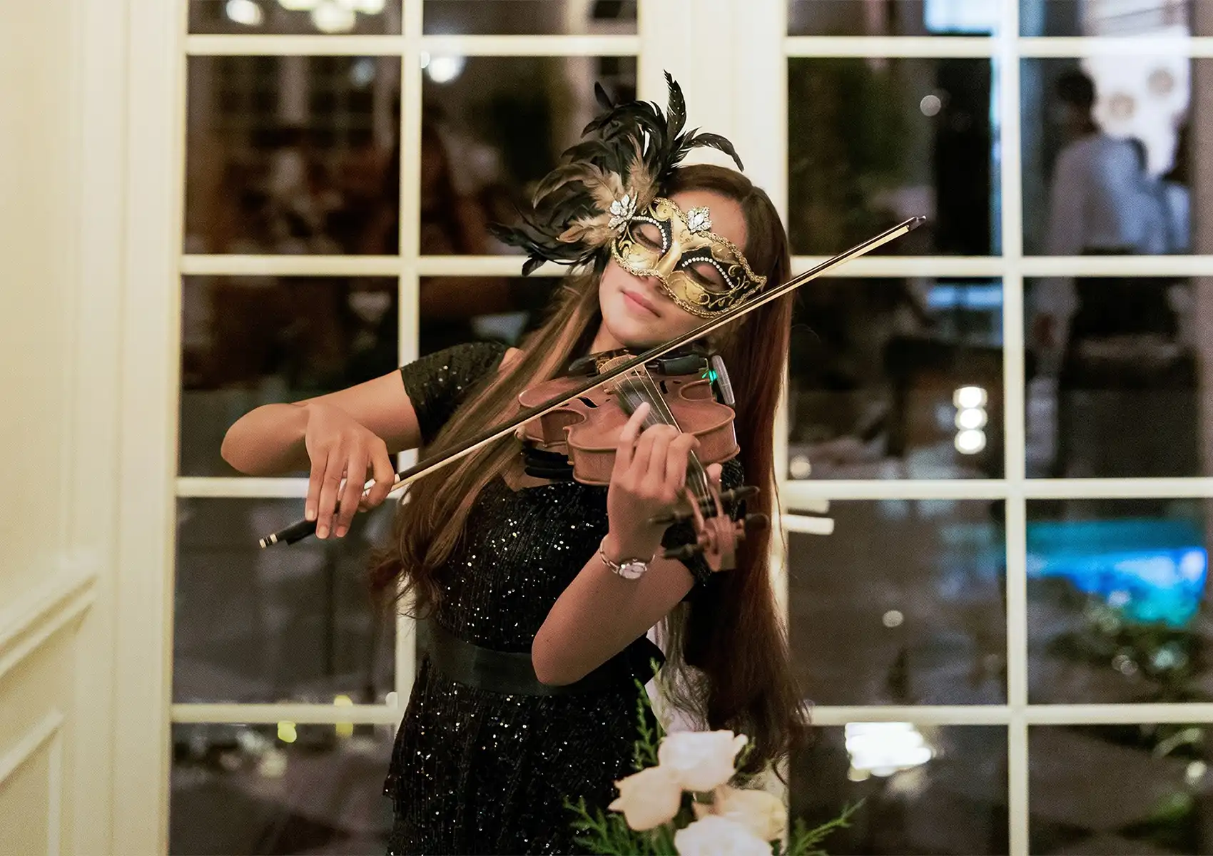 A female violinist wearing a black sequined dress and a gold masquerade mask with feathers performs live music in an elegant room at a boutique hotel bali.