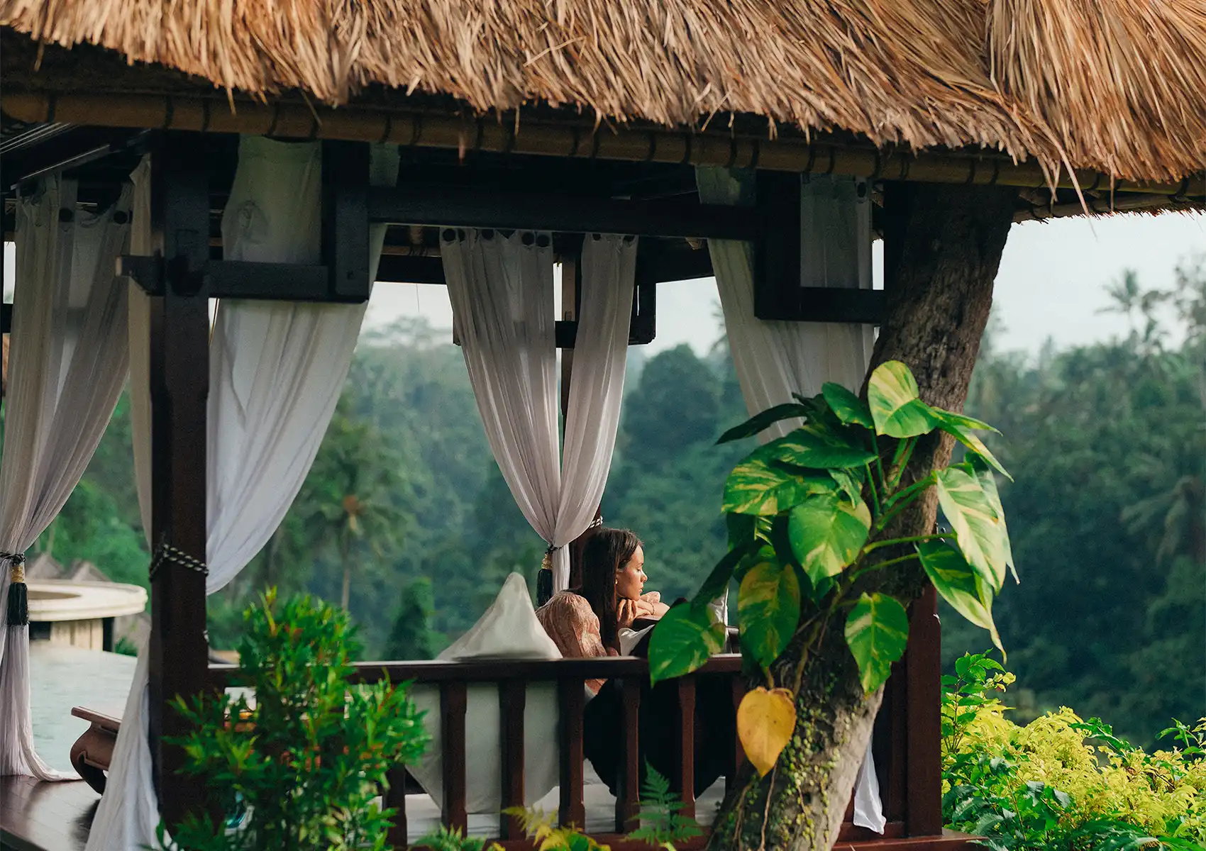 A woman relaxing peacefully inside a traditional thatched-roof gazebo with tied-back white curtains, surrounded by tropical foliage and overlooking a lush green jungle valley at a boutique hotel bali.