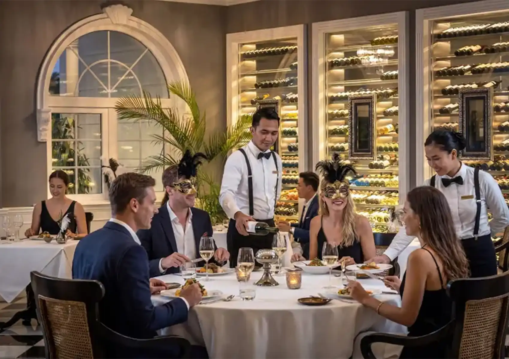 Elegantly dressed guests wearing feathered masquerade masks enjoy a festive dinner while waitstaff pour wine in a fine dining restaurant at a boutique hotel bali.