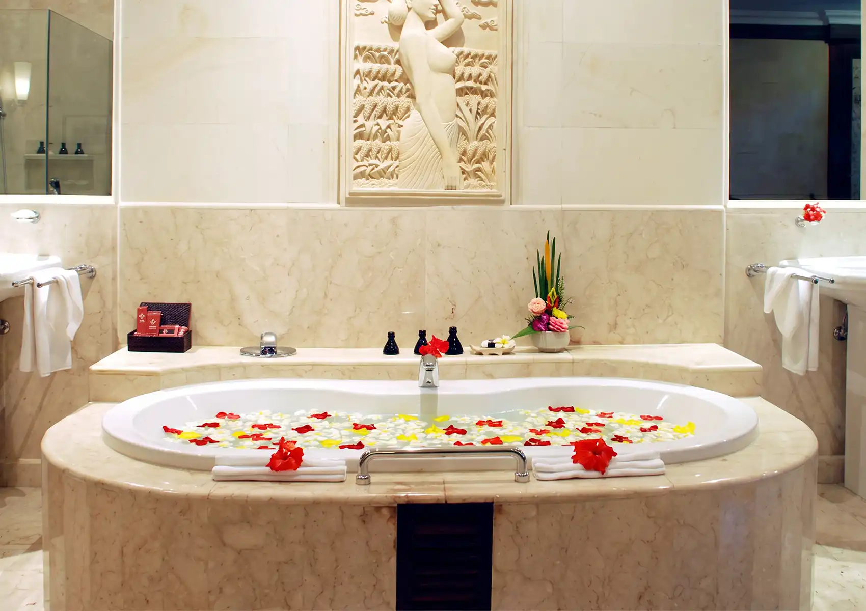 A luxurious marble bathtub filled with red, white, and yellow flower petals positioned below a carved stone wall relief at a boutique hotel bali.