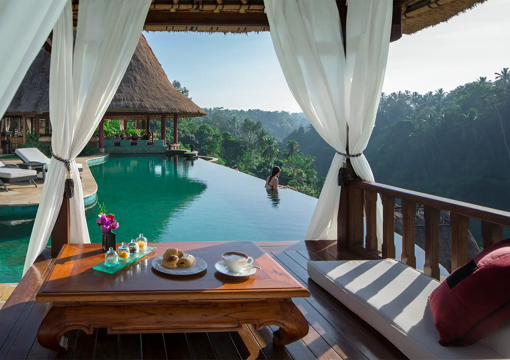 A view from a private thatched-roof pavilion with white curtains, featuring a wooden table with coffee and pastries in the foreground, looking out over an infinity pool and lush jungle valley at a boutique hotel bali.