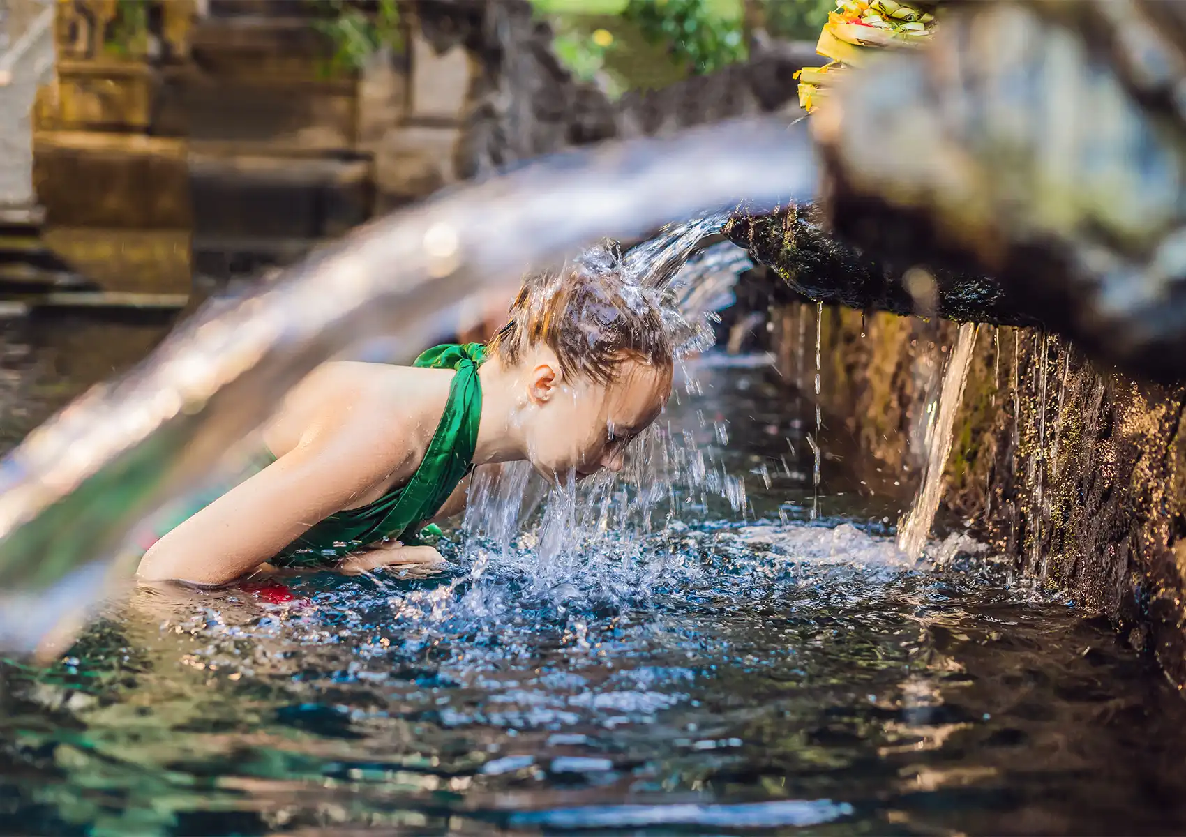 A young woman bowing her head under a flowing water spout in a stone temple pool during a traditional blessing arranged by a boutique hotel bali.
