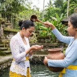 Two women in traditional Balinese dress participating in a sacred water purification ritual with a coconut ladle and clay pot, an excursion offered by a boutique hotel bali.