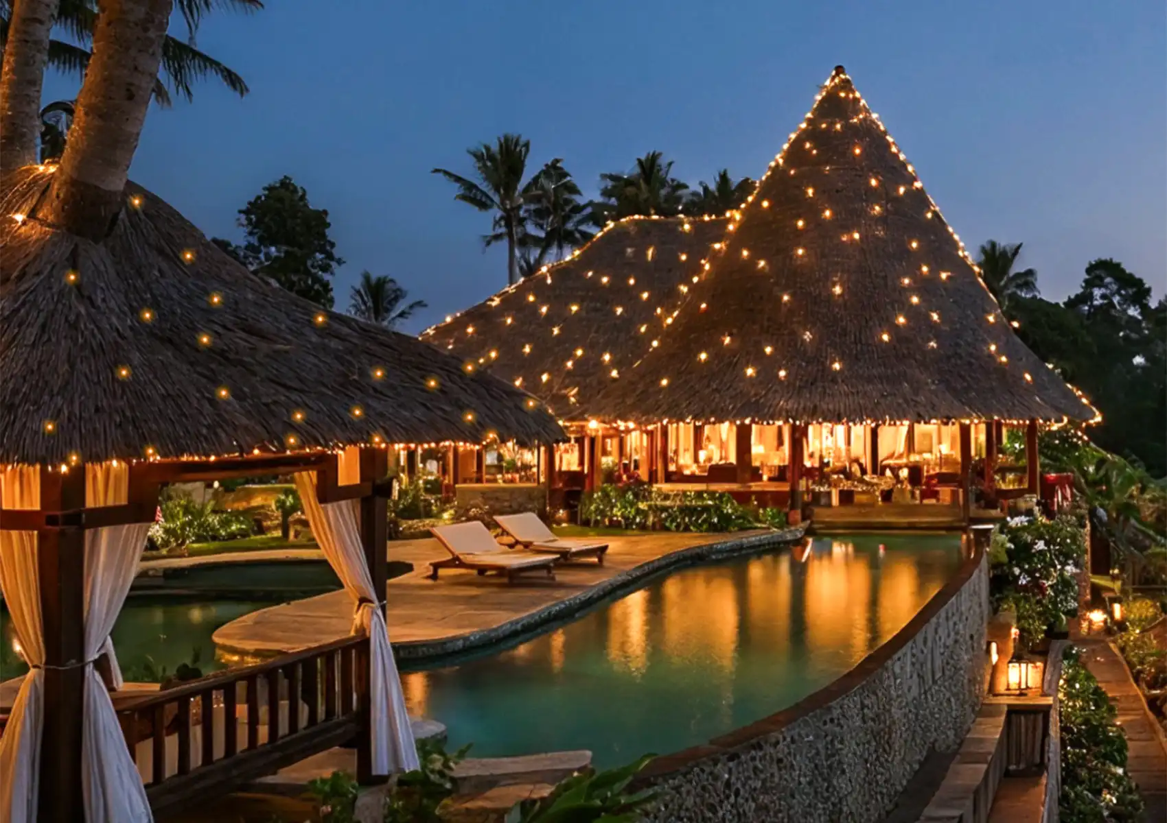Beautifully illuminated thatched-roof buildings decorated with string lights next to a curved swimming pool and lounge chairs at a boutique hotel in Ubud during twilight.
