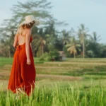 A woman wearing a long, backless red dress and a straw hat walking through a lush green rice paddy field near a boutique hotel Bali, with palm trees and a small hut in the background.