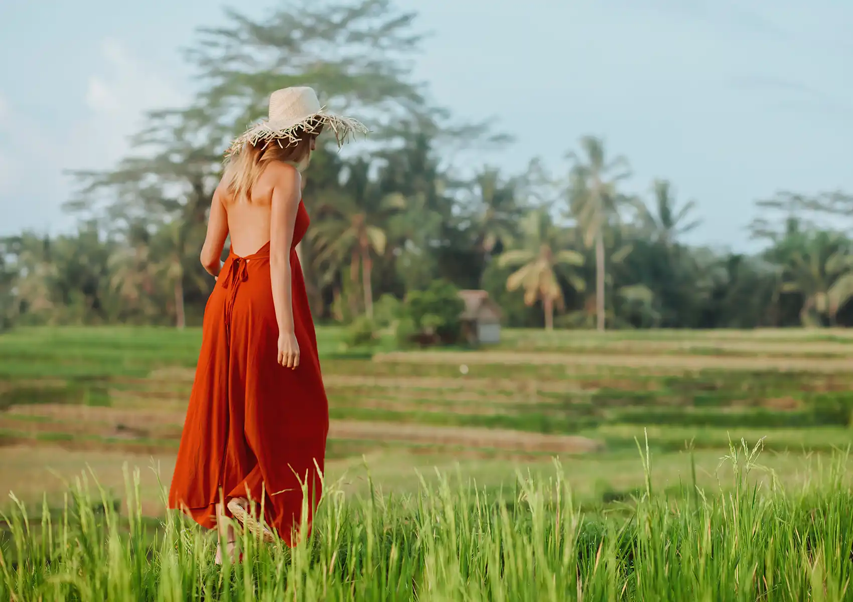 A woman wearing a long, backless red dress and a straw hat walking through a lush green rice paddy field near a boutique hotel Bali, with palm trees and a small hut in the background.