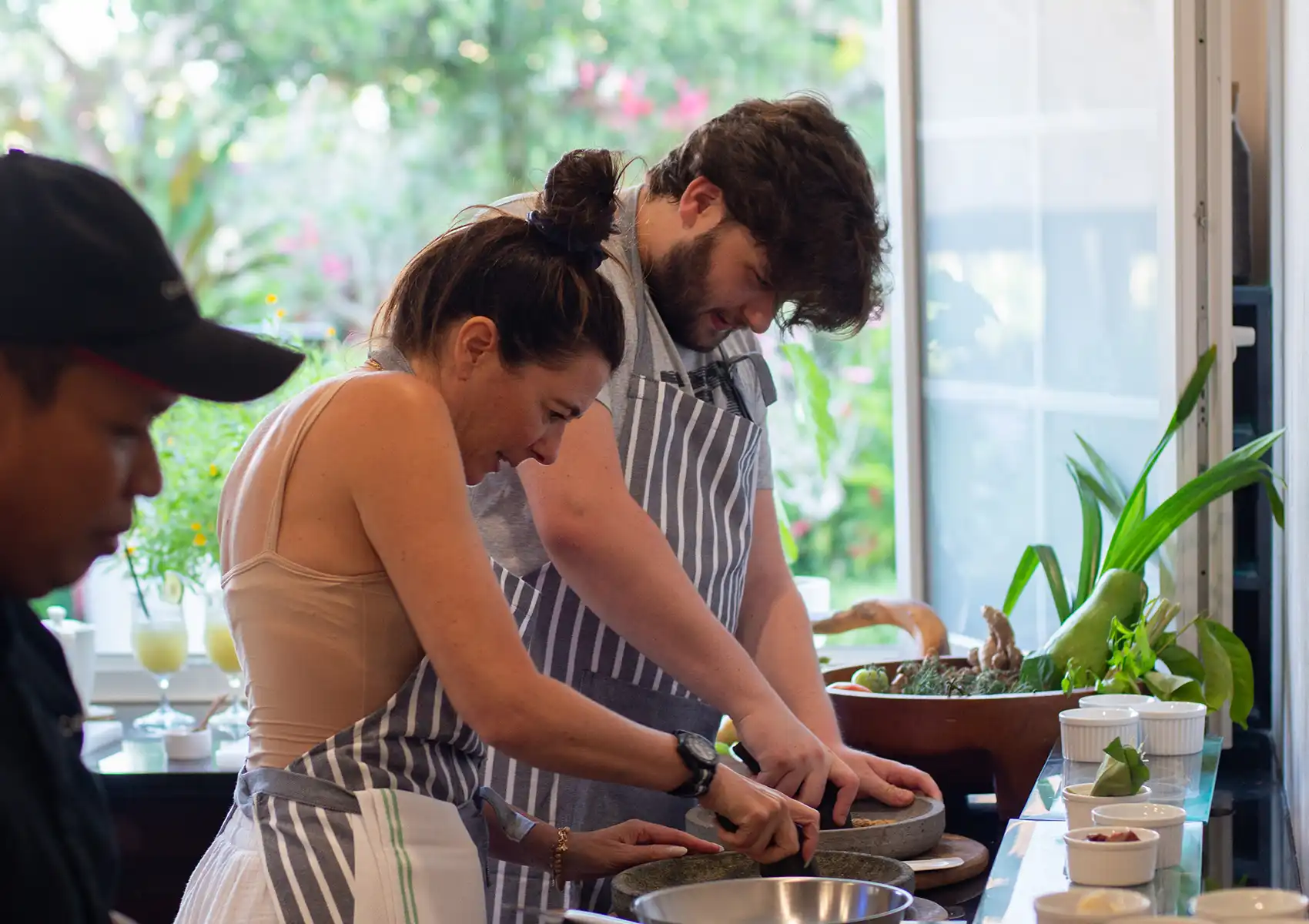 A couple wearing matching striped aprons working together in a bright kitchen, grinding spices with traditional mortars and pestles during a cooking class at a boutique retreat in bali.