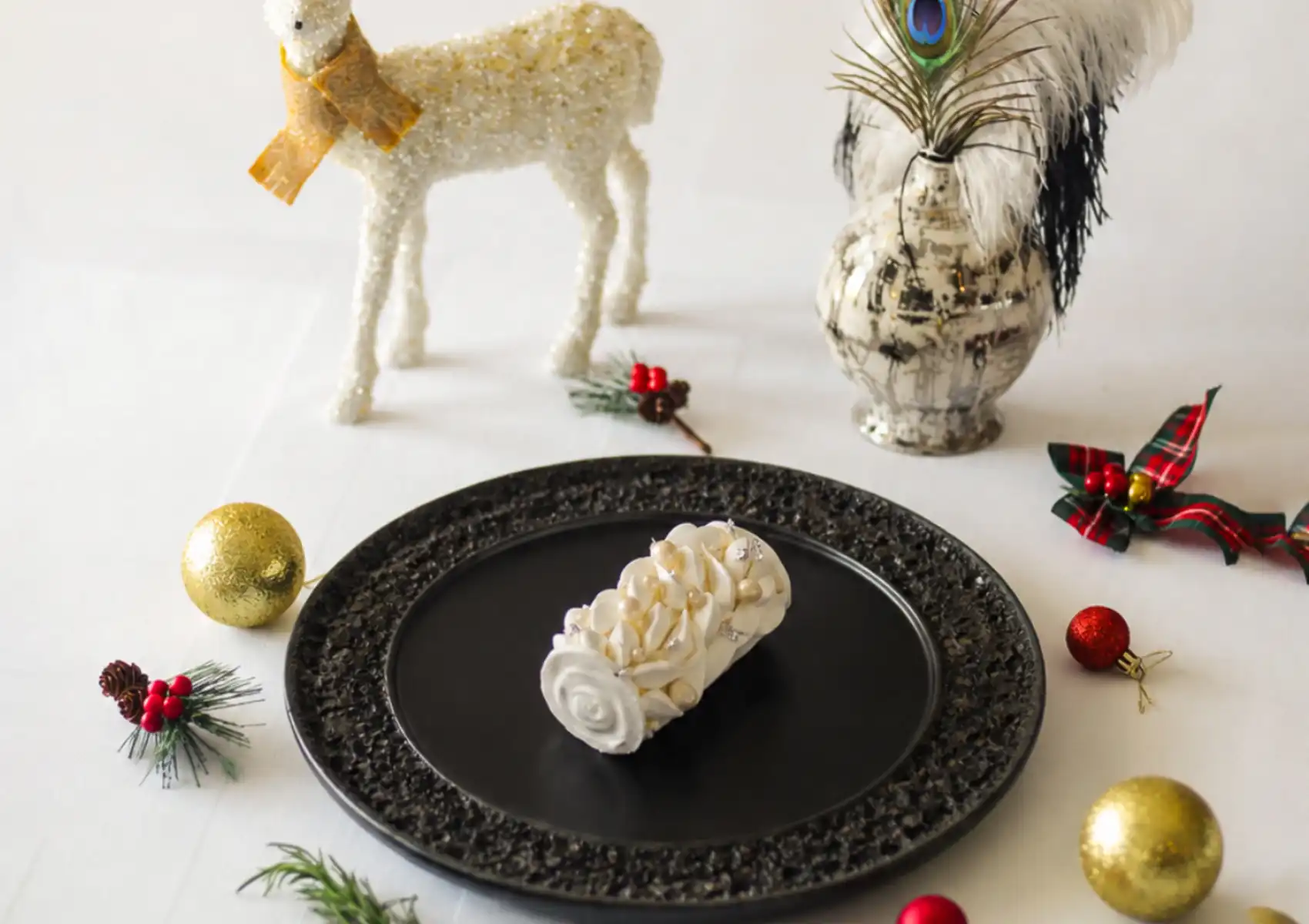 A festive white dessert log served on a textured black plate, surrounded by gold and red ornaments, a glittery reindeer figurine, and peacock feathers to celebrate the holidays at a boutique hotel in Ubud.