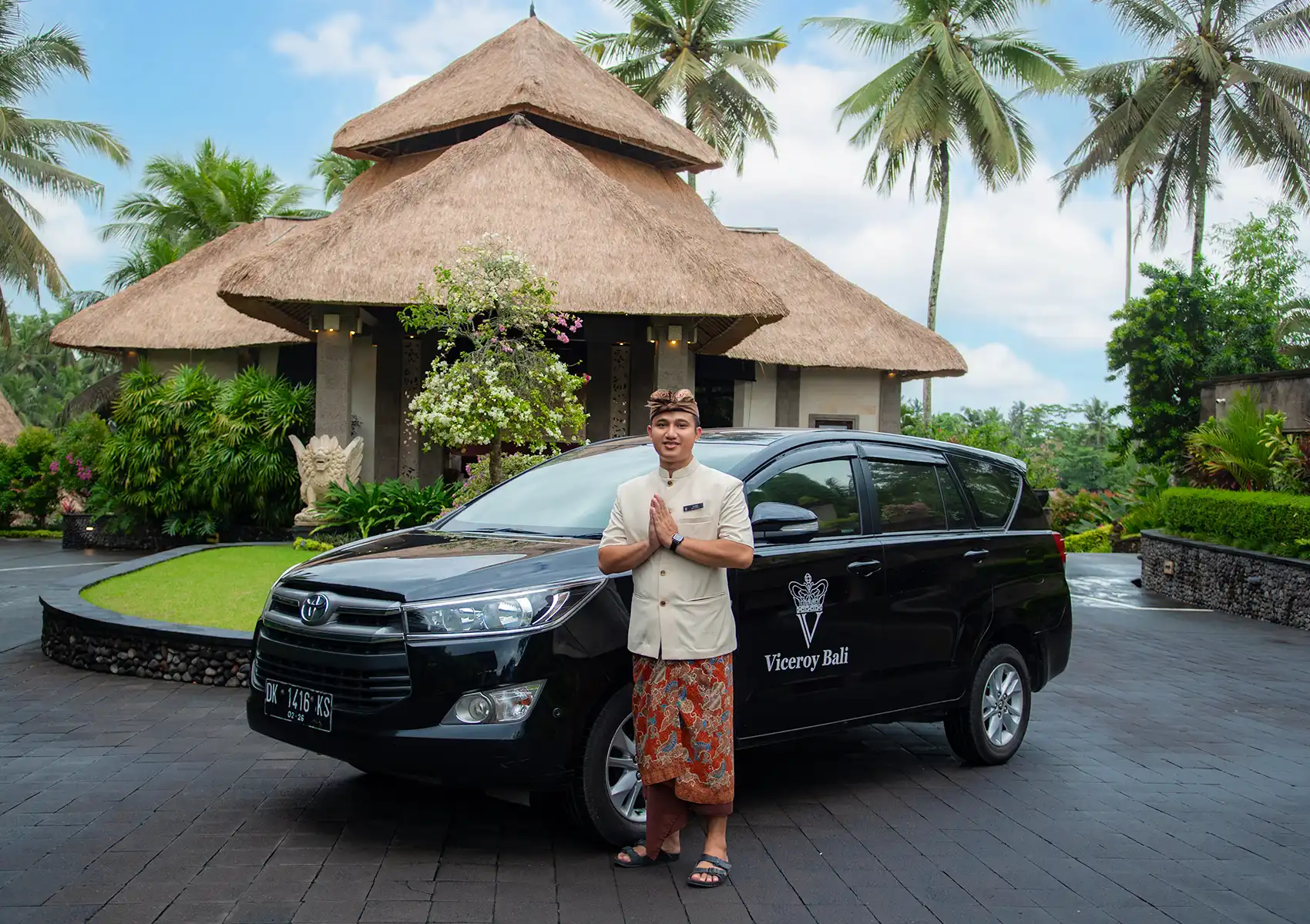 A smiling staff member in traditional Balinese attire greets guests in front of a black Viceroy transfer vehicle at a boutique hotel Bali, featuring a thatched-roof pavilion and palm trees in the background.
