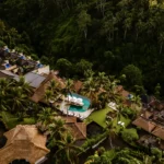 An aerial view of a luxury boutique hotel Bali nestled in a dense jungle valley, showcasing a central infinity pool, palm trees, and terraced thatched-roof villas with private plunge pools.