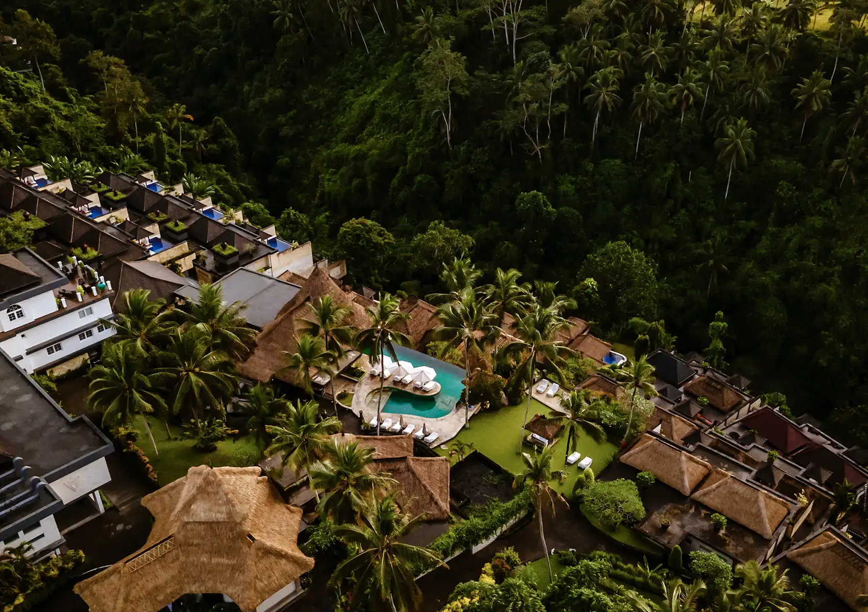 An aerial view of a luxury boutique hotel Bali nestled in a dense jungle valley, showcasing a central infinity pool, palm trees, and terraced thatched-roof villas with private plunge pools.