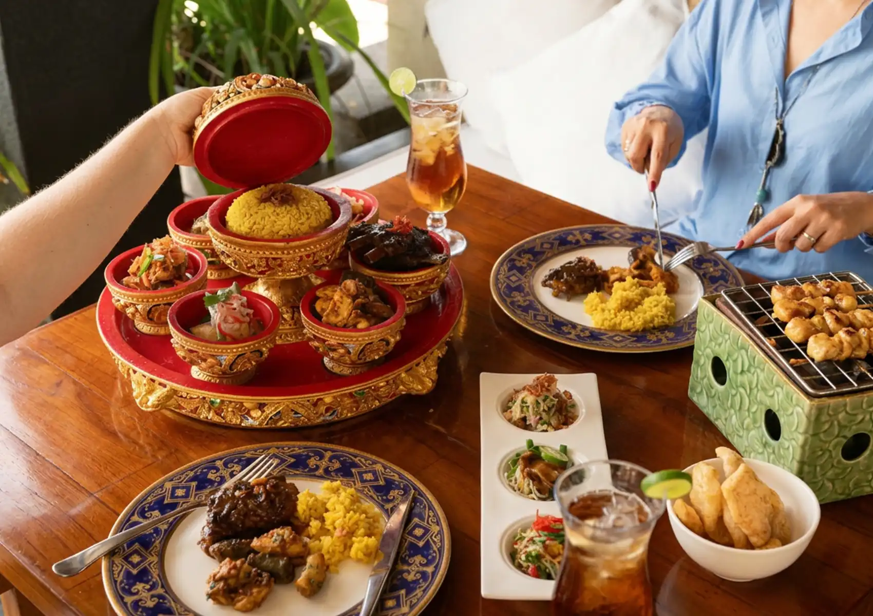 Diners enjoying a shared Balinese feast with traditional dishes served in ornate bowls on a tiered tray, alongside satay on a small grill and iced drinks at a boutique retreat in bali.