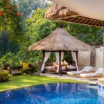 A traditional thatched-roof gazebo and white lounge chairs beside the blue tiled swimming pool of a tropical private pool villa Ubud.