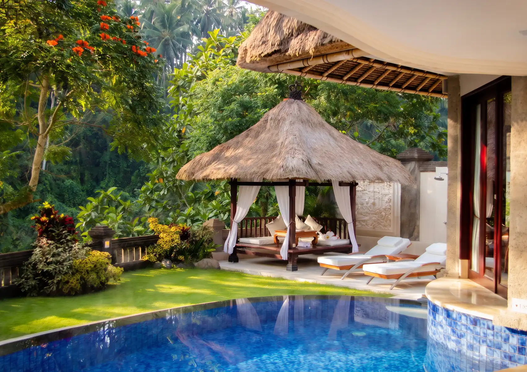 A traditional thatched-roof gazebo and white lounge chairs beside the blue tiled swimming pool of a tropical private pool villa Ubud.